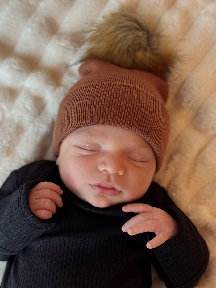 Newborn baby sleeping peacefully in a brown beanie with a fluffy pom-pom, wearing a black long-sleeve outfit.