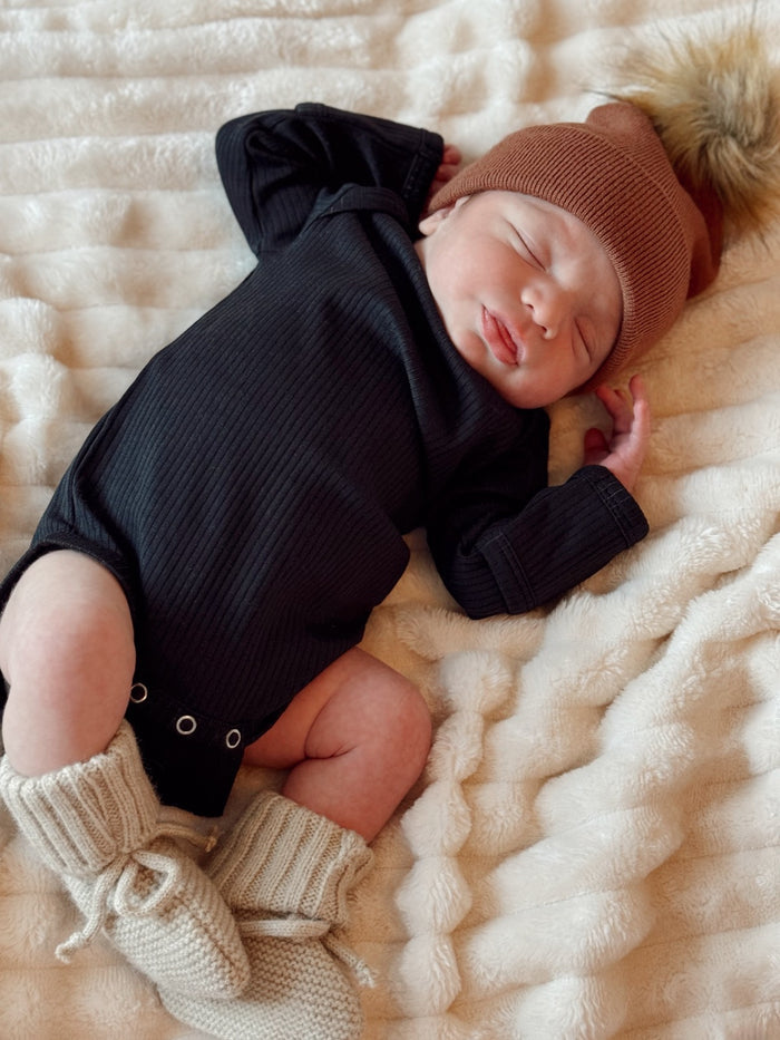 Newborn baby in a black onesie and brown hat, peacefully sleeping on a soft, textured blanket.