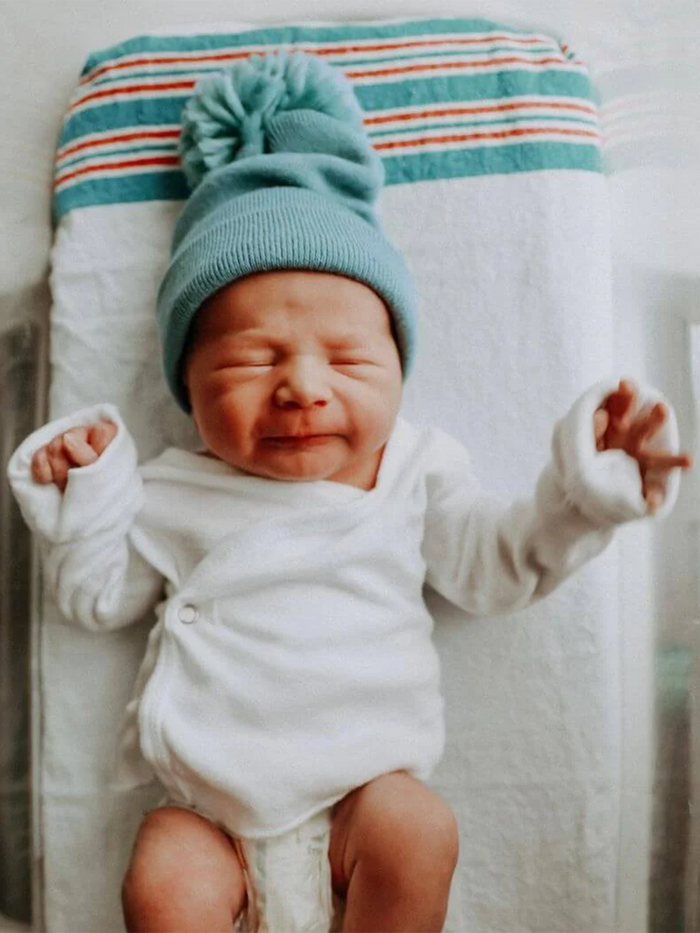 Newborn baby wearing a blue knit hat, smiling while lying on a colorful striped blanket.