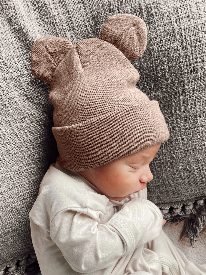 Baby wearing a soft, brown hat with bear ears, sleeping peacefully on a textured gray blanket.