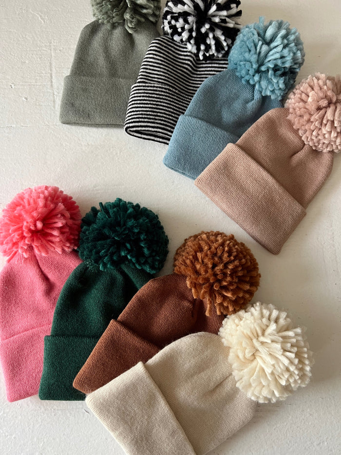 Nine colorful beanies with pom-poms arranged on a light background, showcasing various styles and colors.