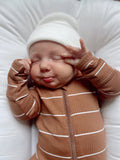Baby with a playful expression, wearing a striped brown outfit and a white hat, sticking out their tongue.