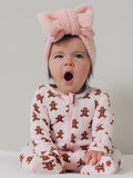 Baby in a pink gingerbread onesie yawns, wearing a fluffy pink headband, sitting against a neutral background.