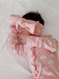 Sleeping baby in a pink outfit with rainbow patterns and a matching headband on a soft white blanket.