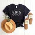 Black t-shirt with "BONUS mama" text, a tan hat, and sandals on a white wooden background.