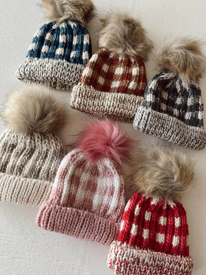 Six knitted beanies with fur pom-poms in various colors and patterns, arranged on a light surface.