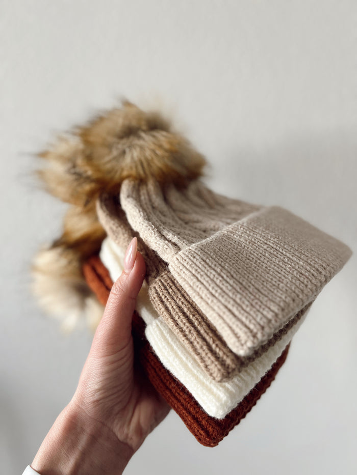 Stack of knitted beanies in beige, brown, cream, and rust colors, held in a hand against a neutral background.