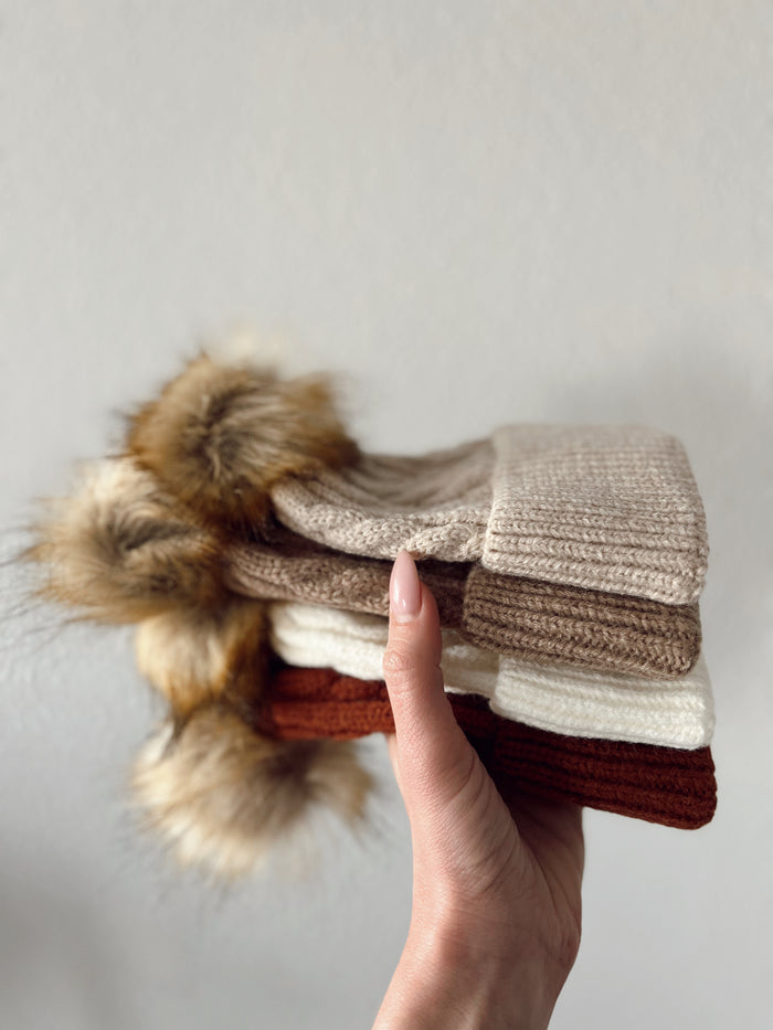 Hand holding a stack of knitted hats in neutral tones with faux fur pom-poms on top.