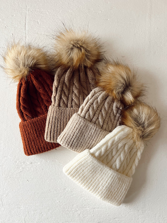 Three knitted beanies in rust, beige, and white colors, each topped with a fluffy pom-pom, on a light background.