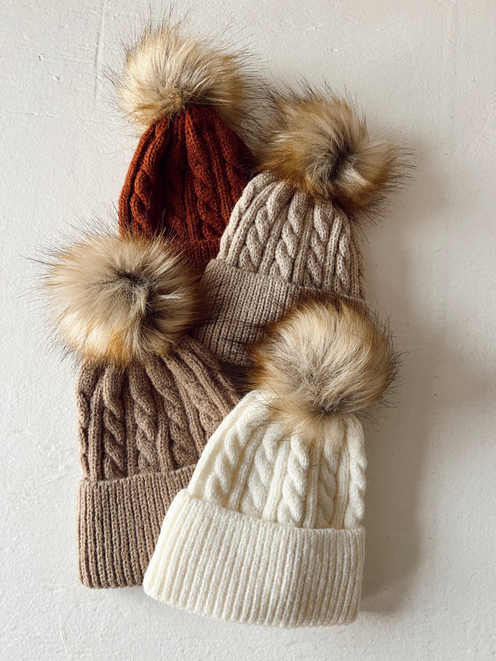 Four knitted beanies in earthy tones with fur pom-poms, arranged on a light background.