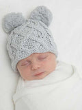 Baby sleeping peacefully, wearing a light gray knitted hat with pom-poms, wrapped in a white blanket.