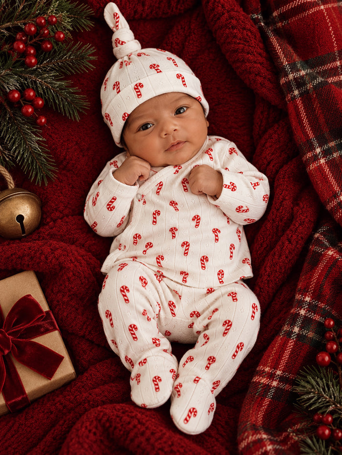 Newborn in candy cane outfit, lying on red blanket with holiday decor and gift box.