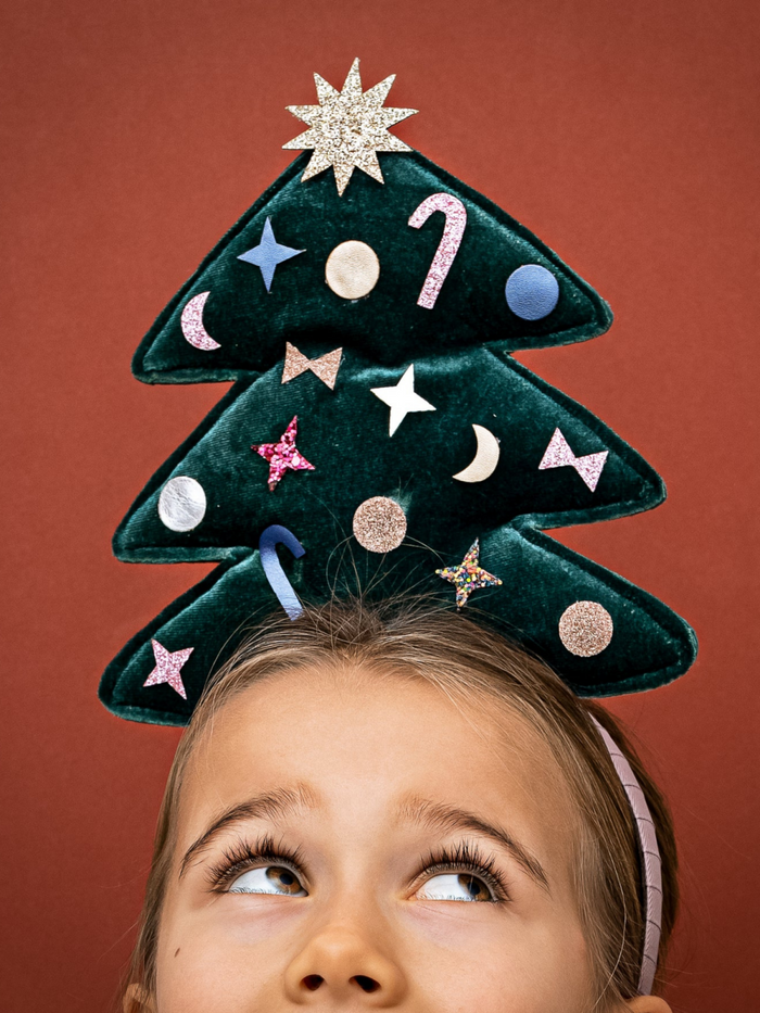Child wearing a festive Christmas tree headband with colorful decorations, looking up against a reddish background.