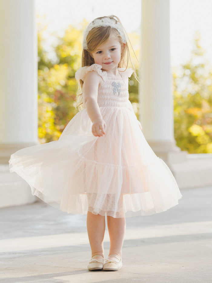 Young girl in a light pink dress twirling, smiling, with a headband, in an outdoor setting with soft sunlight.