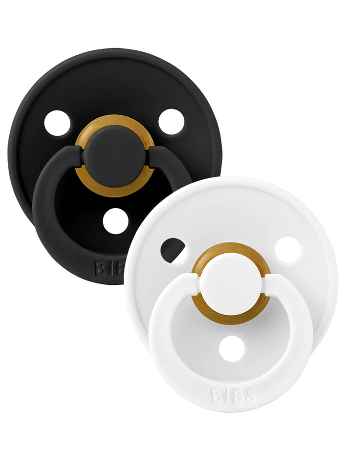 Two pacifiers from BIBS: one black with gold accents, one white with gold accents, placed side by side on a white background.
