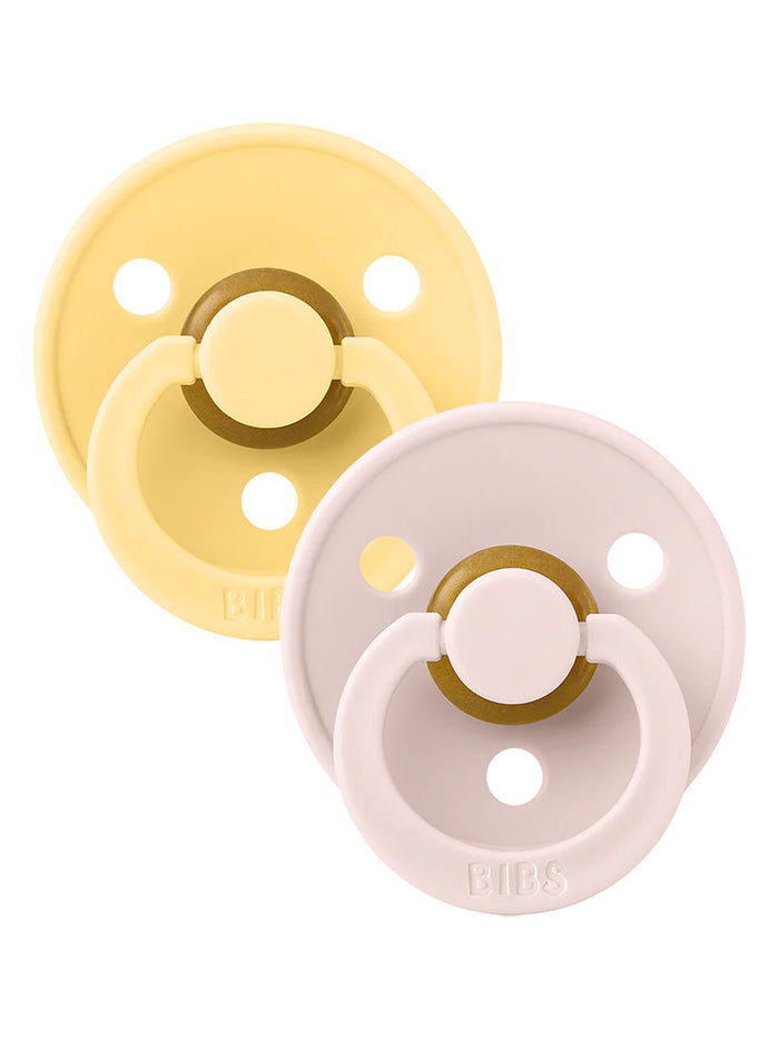 Two silicone pacifiers in soft yellow and pale pink, featuring a round shield and adjustable ring.