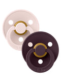 Two pacifiers in beige and dark purple with gold accents, featuring multiple holes for airflow and brand logo.