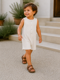 Smiling toddler in a light romper and brown sandals walking on textured outdoor steps. Green plants in the background.