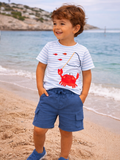 Smiling boy wearing a striped shirt and blue shorts, standing on a beach near water with a crab illustration on his shirt.