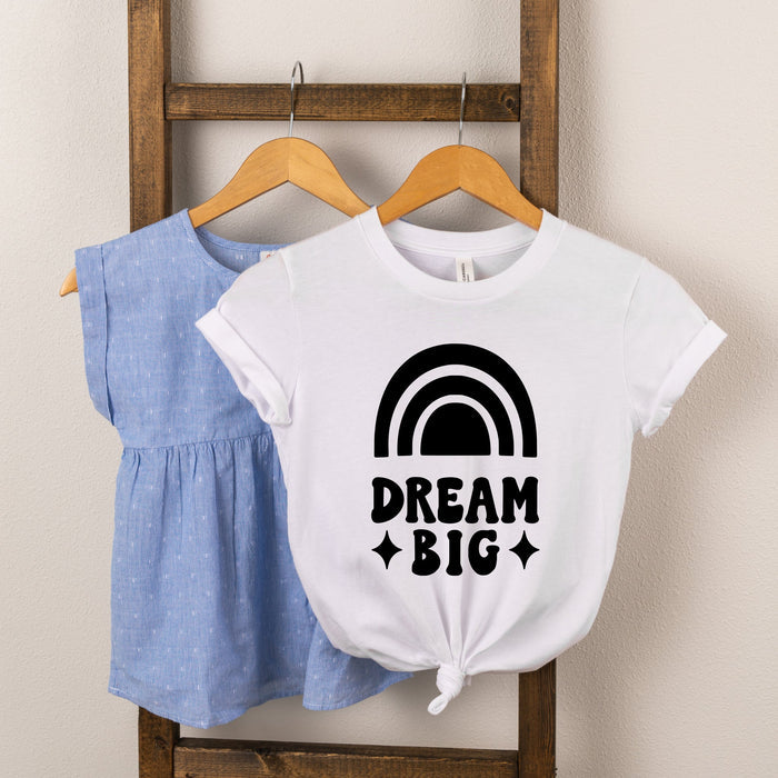 Two children's shirts hanging on a wooden rack: a blue top and a white shirt with a "Dream Big" rainbow design.