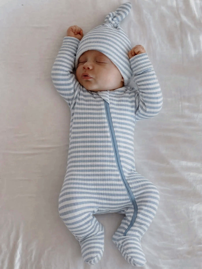 Sleeping baby in a blue striped onesie and matching hat, arms raised, on a soft white surface.