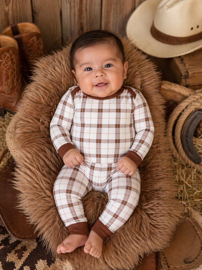 Smiling baby in plaid outfit, sitting on a fluffy rug surrounded by rustic decor, including cowboy boots and a hat.