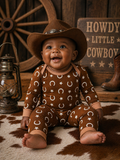 Smiling baby in a cowboy hat, wearing a brown outfit with stars and horseshoes, sitting on a patterned rug.