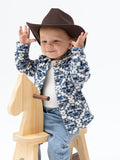Child in a festive sweater and cowboy hat smiles while playing on a wooden rocking horse.