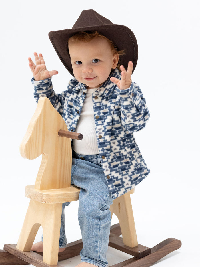 Toddler in a brown cowboy hat waves while sitting on a wooden rocking horse, wearing a patterned sweater and jeans.