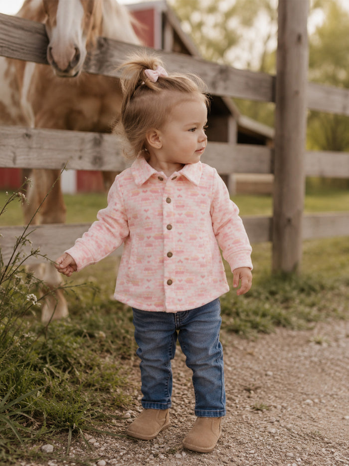 Toddler in a pink shirt and jeans stands by a wooden fence, with a horse in the background.