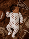 Sleeping baby in a football-patterned onesie, surrounded by footballs and a "Game Day" pennant on a cozy blanket.