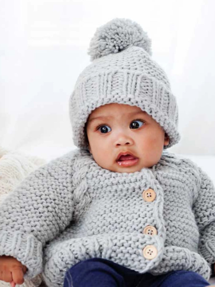 Baby wearing a cozy gray knitted sweater and pom-pom hat, looking curiously at the camera.