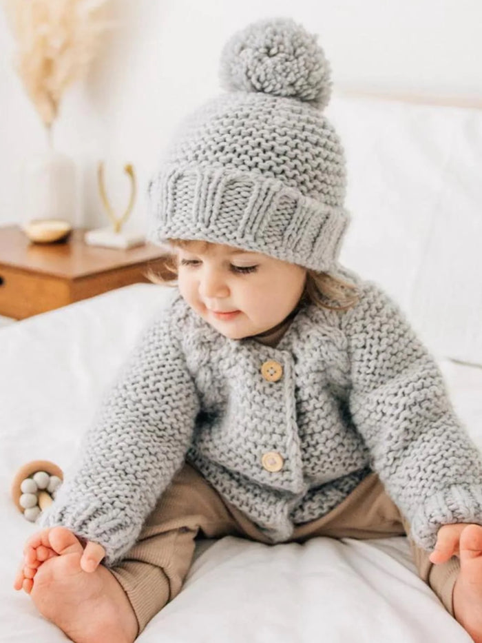 Toddler in a cozy gray knitted cardigan and hat, sitting on a bed with a playful expression.