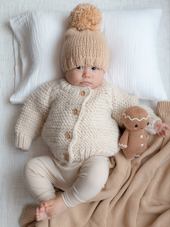 Baby in cozy knitwear and pom-pom hat sits on blanket, with plush toy beside, against a soft backdrop.