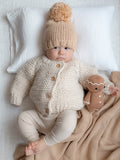 Baby in cozy knitwear and pom-pom hat sits on blanket, with plush toy beside, against a soft backdrop.