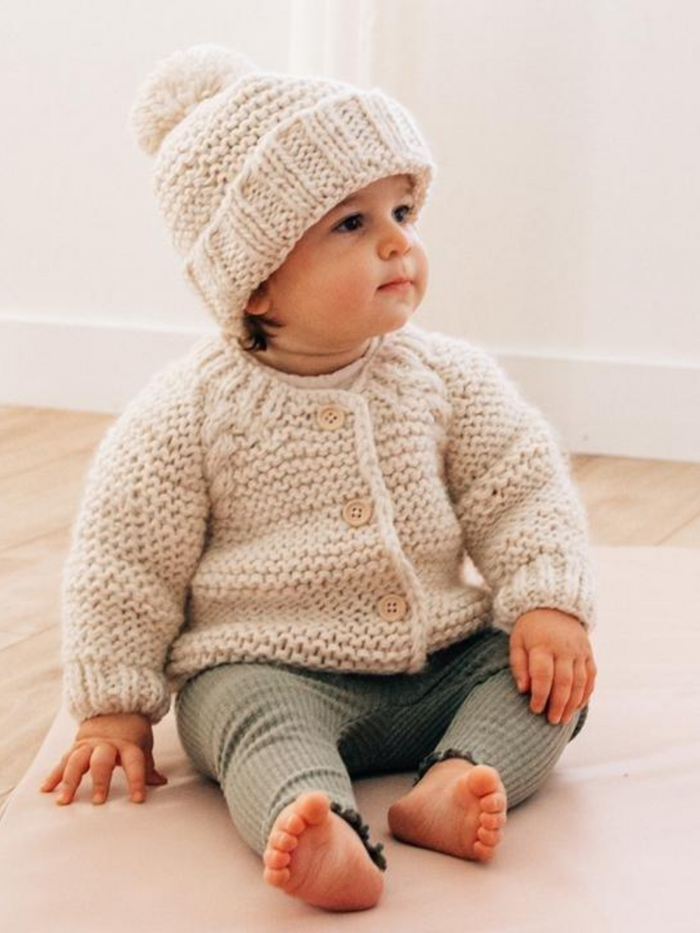 Toddler in cozy cream sweater and hat, sitting on soft surface with wooden floor background.