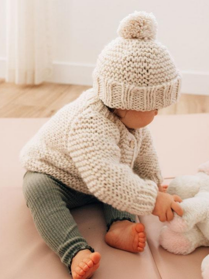 Baby in a knitted sweater and hat sits on the floor, playing with a plush toy. Soft, cozy atmosphere.
