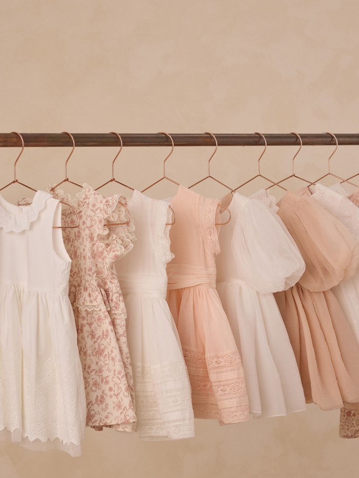 Assortment of children's dresses in soft colors hanging on a copper rack against a beige background.