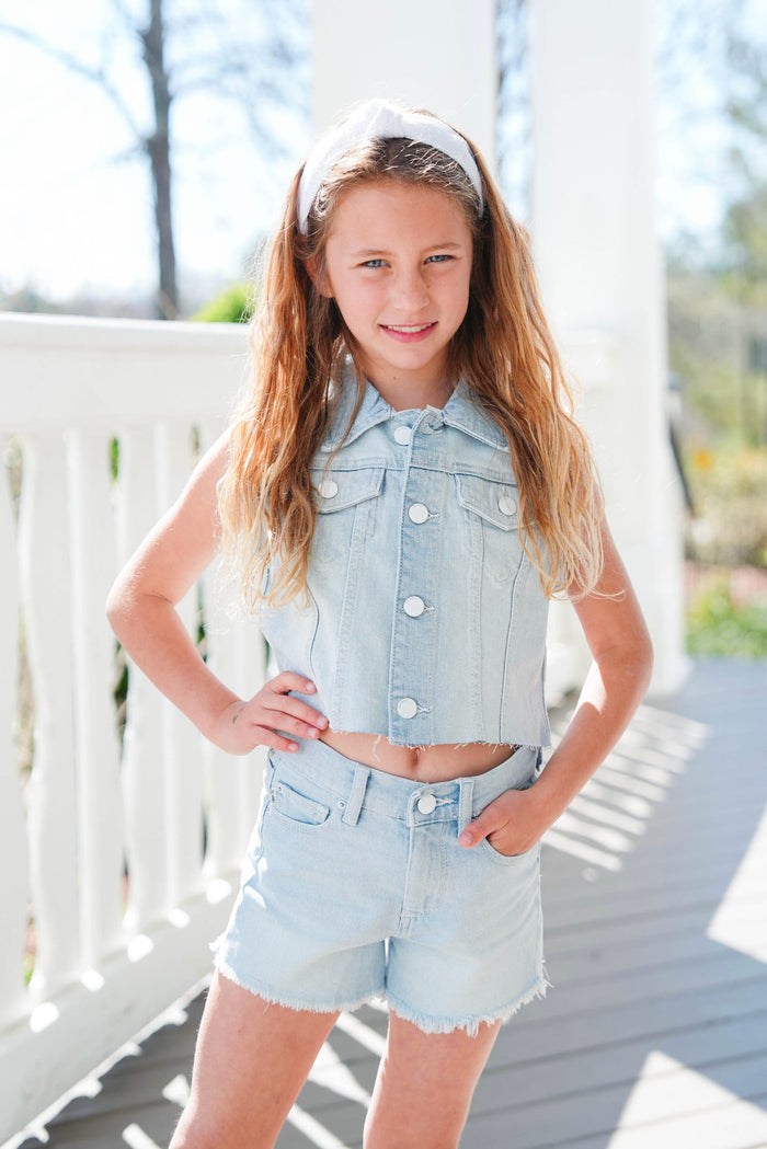 Young girl wearing a denim vest and shorts on a wooden deck.