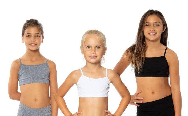 Three girls pose against a white background, wearing gray, white, and black athletic crop tops and bottoms.