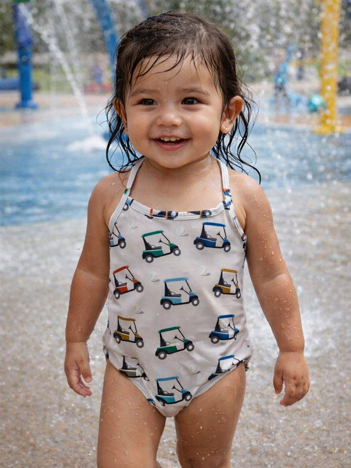 Smiling toddler in a patterned swimsuit standing in a splash pad, with water droplets around them.