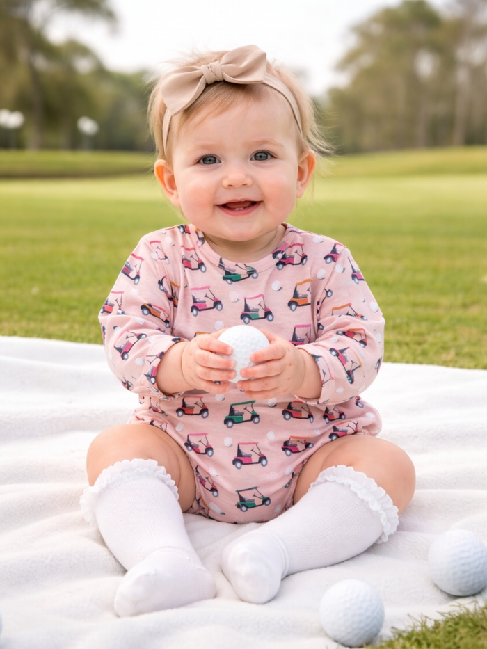 Smiling baby in a pink golf-themed outfit holding a golf ball, sitting on a blanket outdoors.
