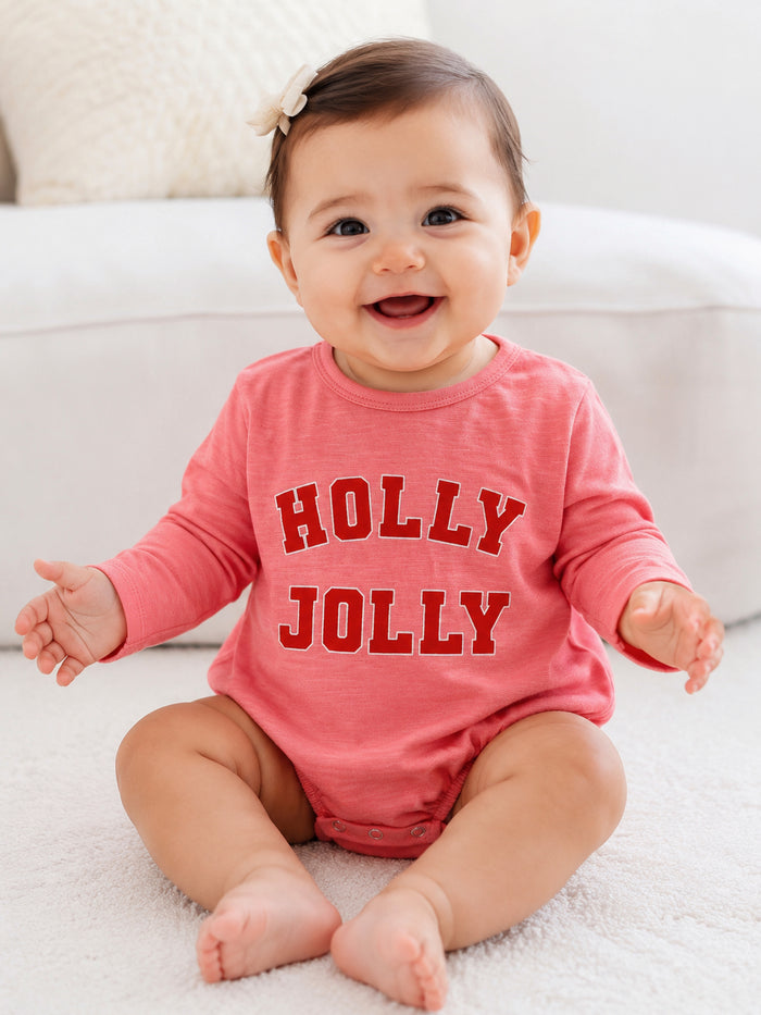 Smiling baby in a pink "HOLLY JOLLY" outfit sitting on a soft surface, with a bow in their hair.