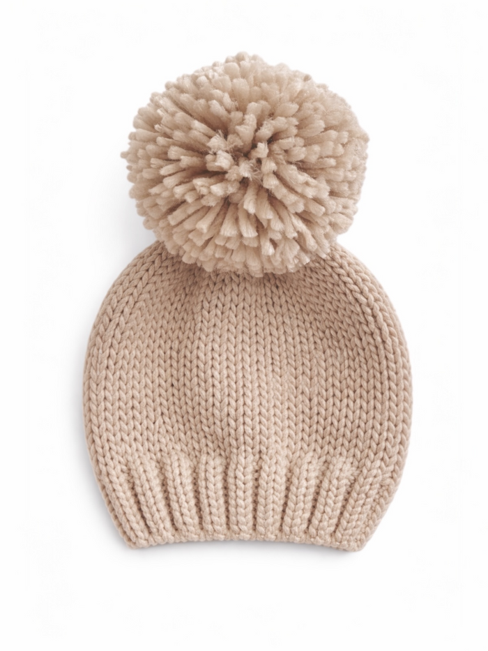 Knitted beige beanie hat with a fluffy pom-pom on top, displayed on a light background.