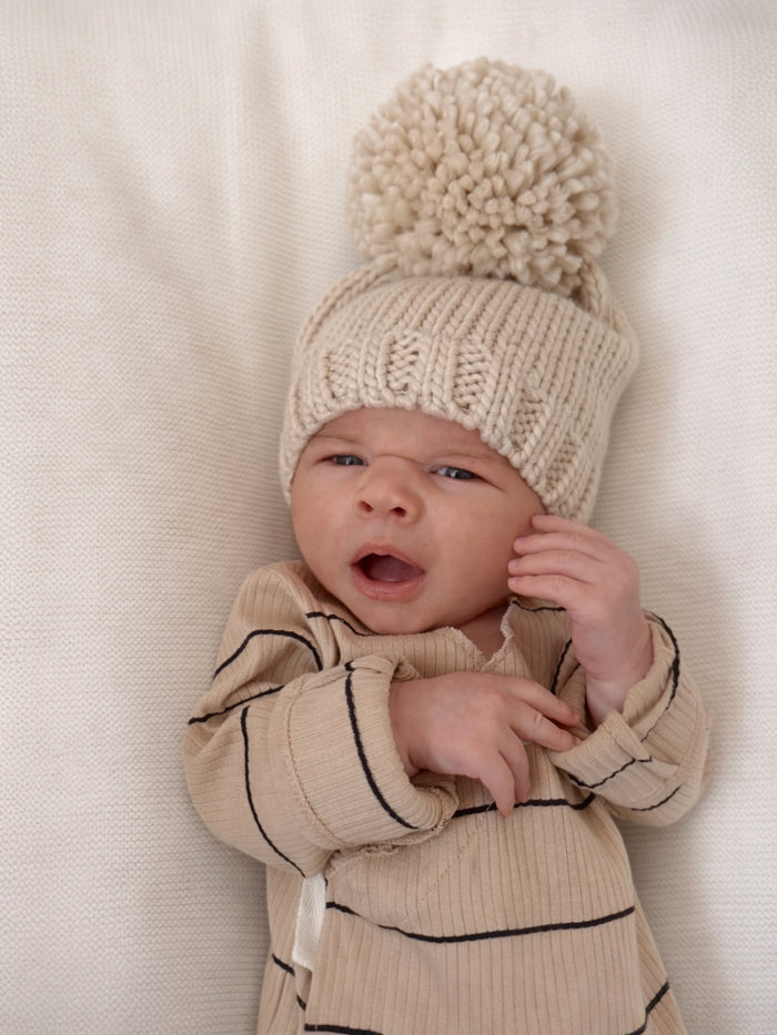 Baby wearing a beige knitted hat with a pom-pom, yawning on a light-colored blanket.