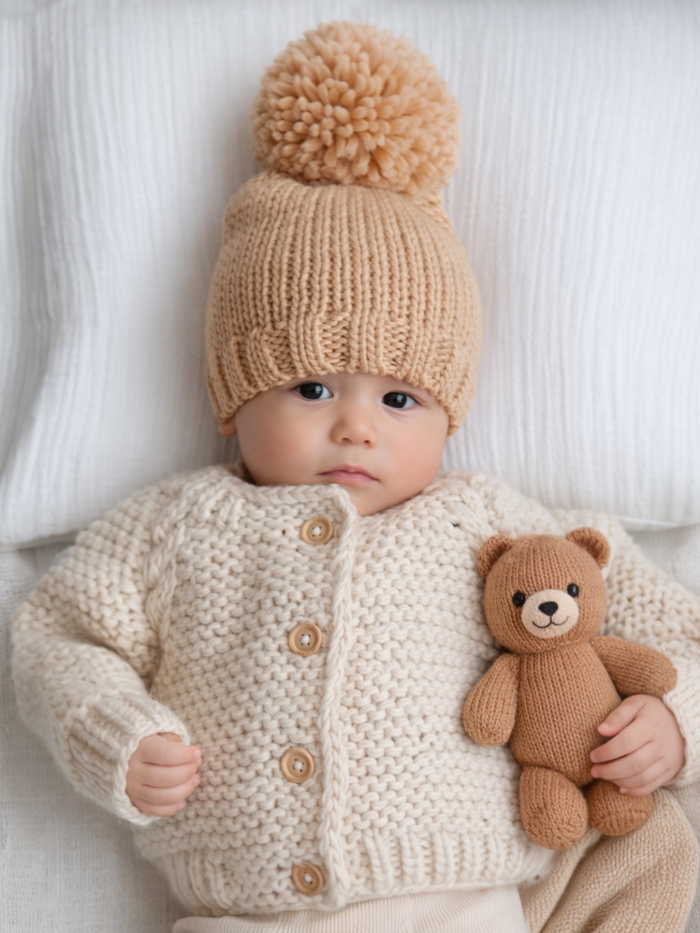 Baby in a cozy cream sweater and tan pom-pom hat, holding a brown teddy bear, lying on a soft surface.