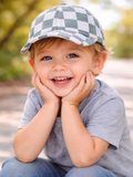 Smiling child with a checkered cap resting chin on hands, sitting outdoors on a path with greenery in the background.