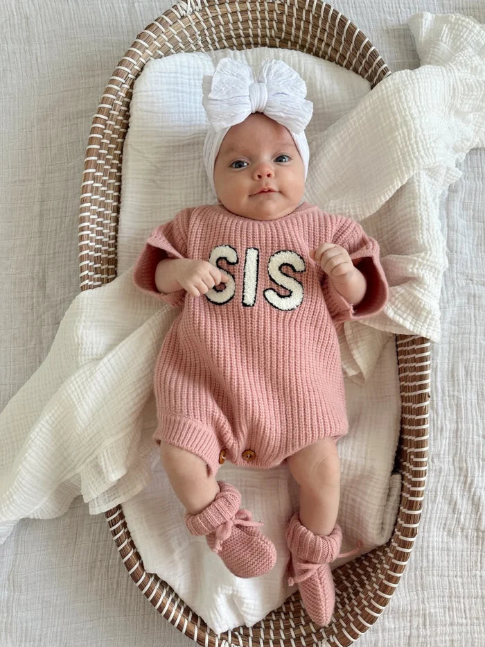 Infant in a pink knitted romper with "SIS" text, wearing a white bow headband, resting in a woven basket.