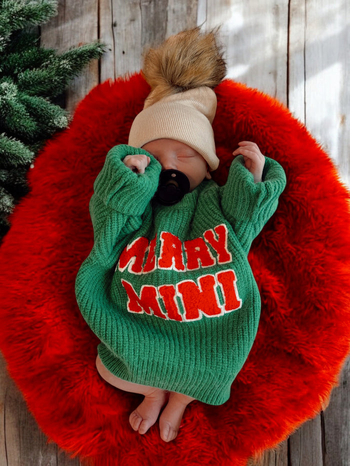 Newborn in a green sweater reading “Merry Mini,” resting on a red fuzzy blanket with a playful tan hat.
