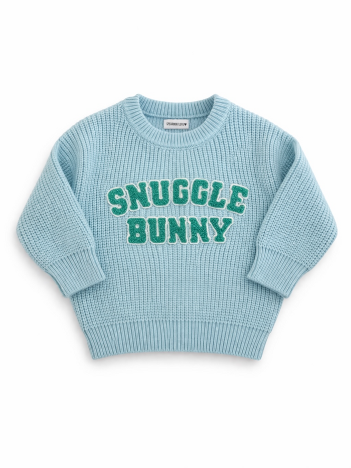Light blue knit sweater with "Snuggle Bunny" in green text on the front. Cozy and stylish for children.